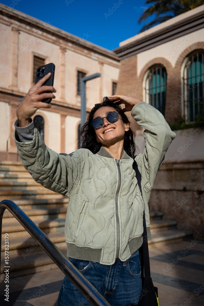 Fototapeta premium Stylish young woman in sunglasses taking selfie with smartphone on sunny day in urban setting.