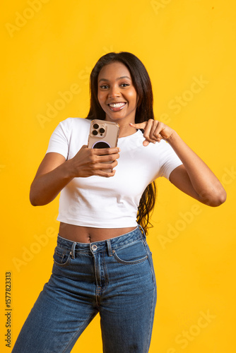 Smiling model pointing at smartphone on yellow background