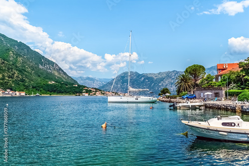 Sailboats docked along the serene waters of Boka Kotorska Bay in Kotor, Montenegro. Picturesque landscape with mountains and coastal houses under a clear sky