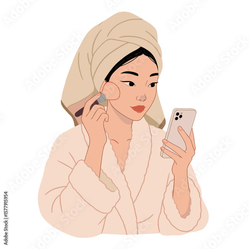 Woman in bathrobe and towel using phone while applying makeup with a brush on her face in a relaxed setting