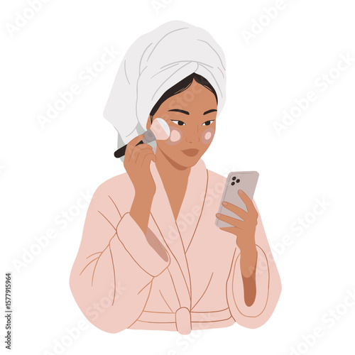 Woman in robe and towel applying makeup while looking at her phone in a self care beauty routine