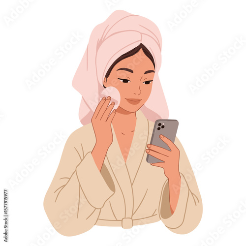 Woman in robe and towel using phone while applying skincare with cotton pad in a self care routine