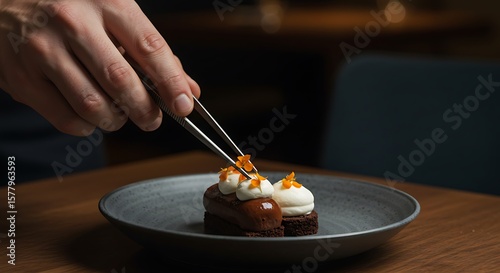 Chef is using tweezers to decorate a dessert with orange flowers on a dark plate on a wooden table