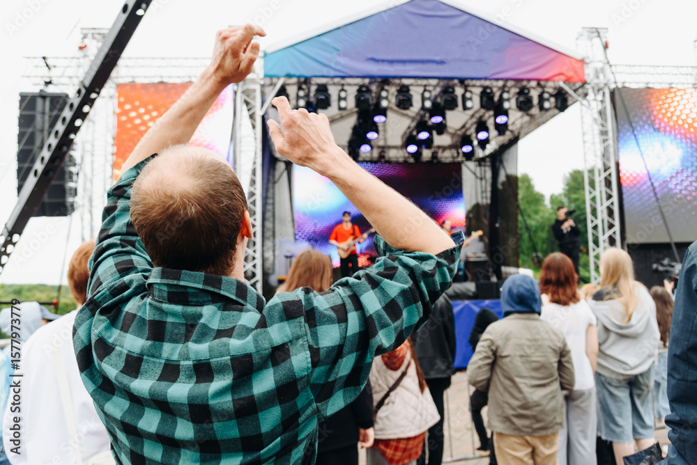 Obraz premium music festival in city park, crowd of people backview, rock group performing on stage, green trees and summer day