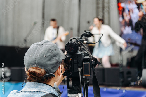 large black video camera on tripod, close-up view, videographer working at music festival in city park, rock group performing on stage