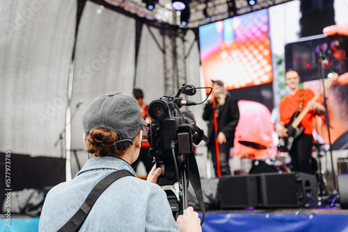 large black video camera on tripod, videographer working at music festival in city park, rock group performing on stage