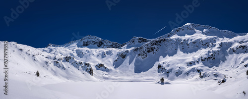 Frozen Ribno lake covered by deep snow against Pirin mountains in Bulgaria near Bansko. White snowy mountains under the clear blue sky in sunny day.