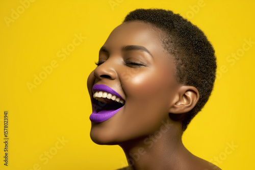 A portrait of a beautiful African American woman with short hair and purple lipstick on her lips laughing out loud, yellow background. 