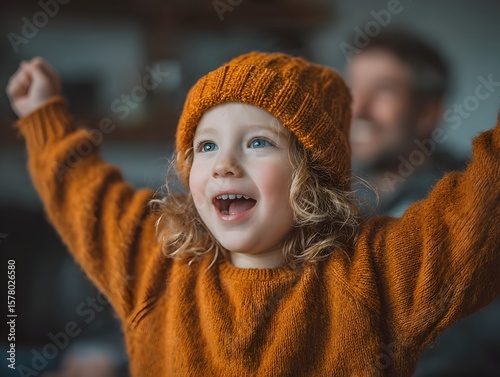 An elated toddler wea an orange hat and sweater joyfully raises their arms, celebrating a fun day indoors.