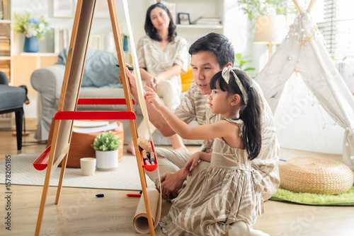 Happy Asian family spending quality time together at home. Father and daughter drawing on the floor with colored pens while mother works on a digital tablet smartphone on the sofa. Warm, modern living