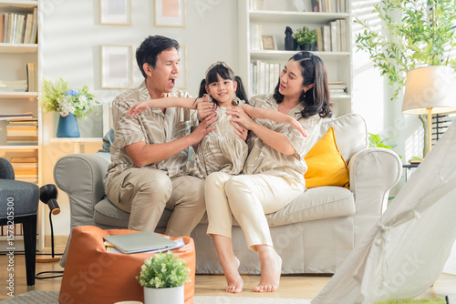 Happy Asian family enjoying quality time in their modern living room. Cheerful young parents playfully carry and bond with their daughter while laughing together on the sofa, creating joyful moments 