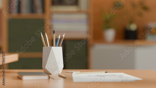 A 3D-rendered geometric white pencil holder with angular faceted design sits on a wooden desk, containing colored pencils and pens, surrounded by notebooks and office supplies in natural light.