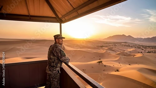 Soldier in camouflage gazing out at desert sand dunes from observation point during warm sunset with mountainous background