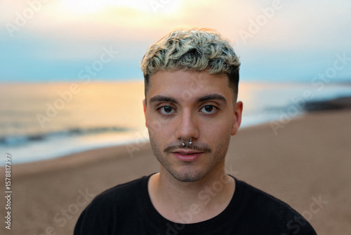 Young man with bleached curly hair and a septum piercing standing confidently on a beach at sunset, expressing individuality and modern style.
