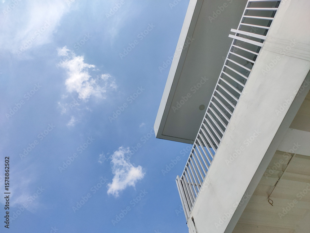 Fototapeta premium A dynamic low angle view shows the modern architectural details of a building with a clean white balcony railing under a vast blue sky with scattered white clouds.
