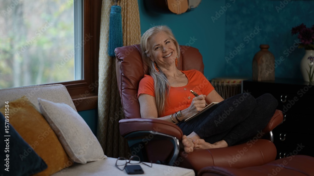Φωτογραφία Portrait of writing in a journal notebook with a pen, legs crossed and barefoot a mature woman with long blonde hair sits in a brown leather recliner, wearing an orange shirt and glasses