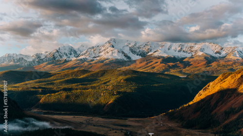 Golden hour sunlight illuminates a vast, verdant valley nestled beneath towering, snow-capped peaks, creating a breathtaking panorama of nature's grandeur