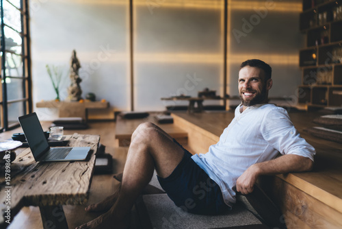 Smiling freelancer reclining in relaxed posture beside laptop, portraying harmony between professional focus and emotional wellbeing in independent tech work lifestyle within natural space.
