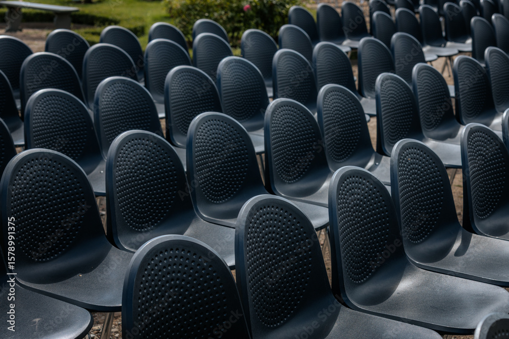 Obraz premium Multiple rows of identical black chairs are neatly arranged in an outdoor setting on a dirt surface, creating a uniform and symmetrical pattern.
