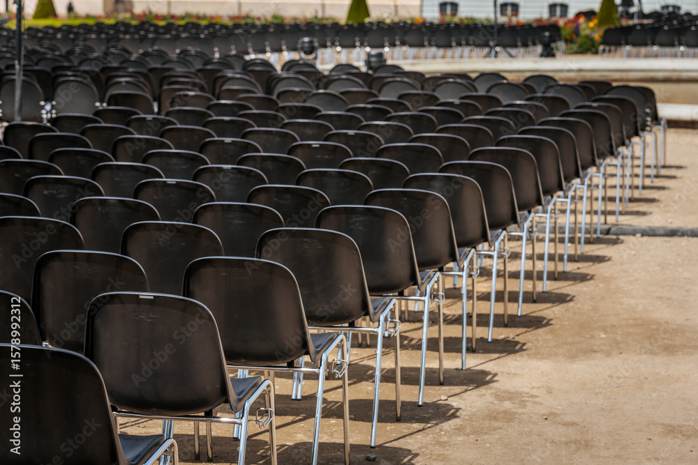 Fototapeta premium Multiple rows of identical black chairs are neatly arranged in an outdoor setting on a dirt surface, creating a uniform and symmetrical pattern.