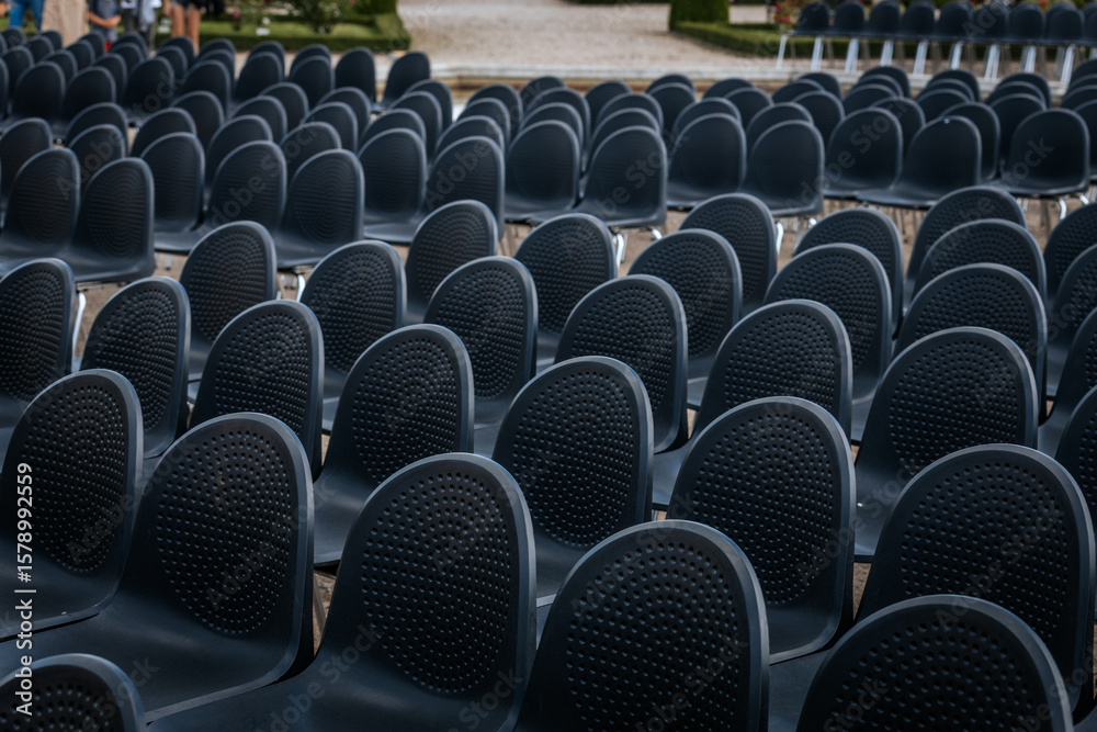 Obraz premium Multiple rows of identical black chairs are neatly arranged in an outdoor setting on a dirt surface, creating a uniform and symmetrical pattern.