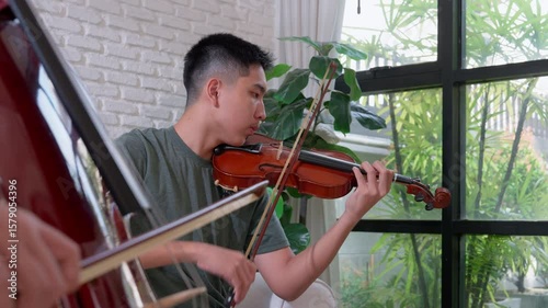 Asian young men practice violin on sofa, their skilled hands guiding the bow across the strings to produce melodious tunes, artistic lifestyle playing violin in living room for practice  and  hobby