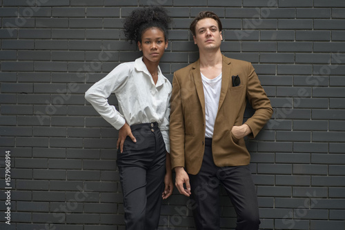 Confident young man and woman in smart casual attire standing against a modern dark brick wall