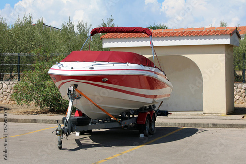 A sleek red and white fiberglass speedboat is tightly strapped to a double-axle trailer, parked in a sunlit lot beside a stucco shelter, ready for transport or storage