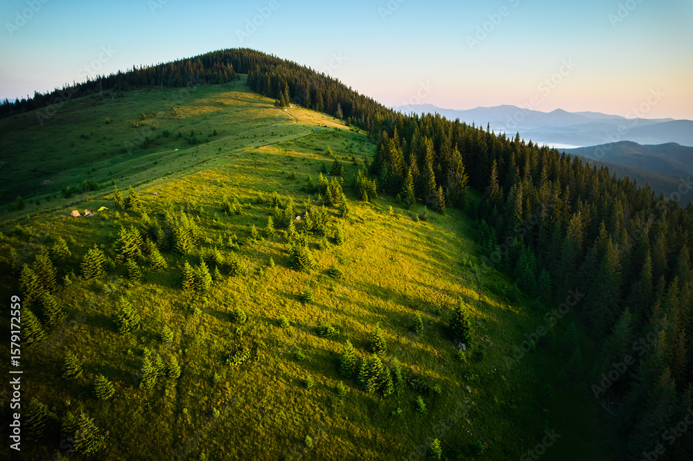 Obraz premium Aerial view of campsite on grassy hillside, with tents scattered amidst lush greenery and small trees. Golden hues of sunrise. In distance, layers of rolling mountains fade into horizon.
