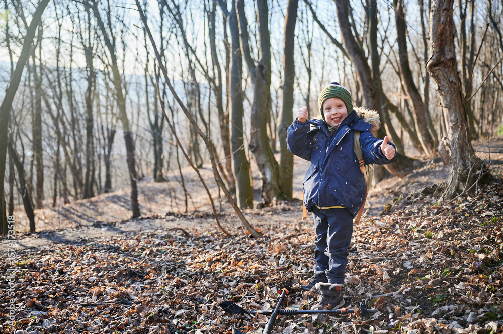 Naklejka premium Young child hiking through forest in early spring, warmly dressed in jacket with fur-lined hood, green beanie. Boy holding trekking poles, exploring leaf-covered forest floor surrounded by tall trees.