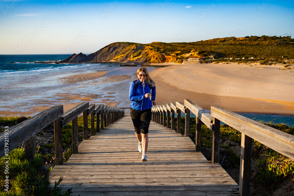Fototapeta premium Beautiful mid adult woman running up wooden stairs at viewpoint on Atlantic ocean and Amoreira beach in Portugal on spring evening. Front view 