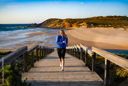 Beautiful mid adult woman running up wooden stairs at viewpoint on Atlantic ocean and 
Amoreira beach in Portugal on spring evening. Front view	