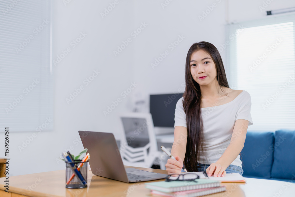 Young Asian businesswoman working from her home office, sitting at a desk, taking notes on a notepad while surrounded by a laptop and various stationery items on the table Young Asian businesswoman working from her home office, sitting at a desk, taking notes on a notepad while surrounded by a laptop and various stationery items on the table