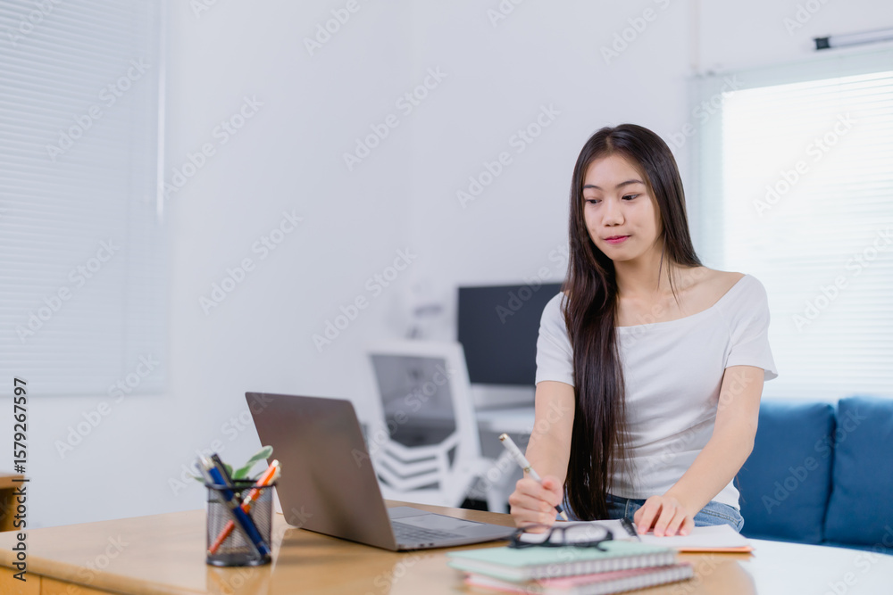 Focused Asian businesswoman sitting at a desk, taking notes and working on a laptop in a home office, embodying remote work and a flexible freelance lifestyle Focused Asian businesswoman sitting at a desk, taking notes and working on a laptop in a home office, embodying remote work and a flexible freelance lifestyle
