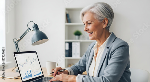 Smiling mature woman working on a laptop, analyzing financial charts in a bright, modern office setting, displaying expertise.