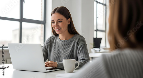 A young woman smiles as she works on her laptop at the office, enjoying a coffee break during her busy work day.