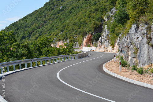 A winding panoramic road above Veli Dol curves through a lush green mountainous landscape with rugged rock faces, featuring a protective guardrail and clear white road markings under a bright sky