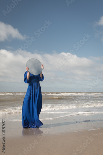 woman in long blue dress on the beach standing in shore holdint roud mirror hiding her face