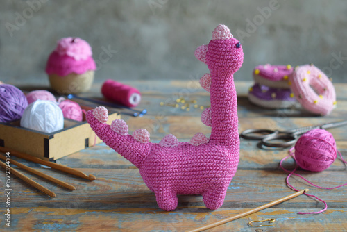 Close up of finished pink amigurumi dinosaur toy on rustic painted wooden table with wool yarn accessories and colorful thread balls crochet craft concept