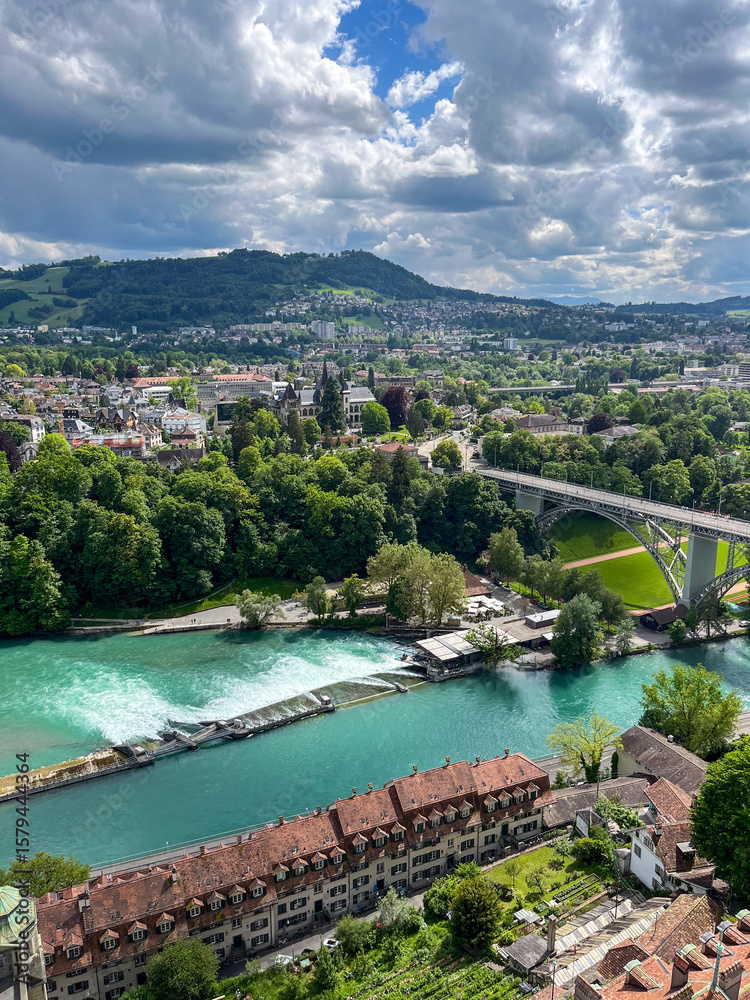 Naklejka premium Scenic view of a vibrant river flowing through lush green landscapes, with bridges connecting urban areas and dramatic clouds creating an atmospheric backdrop