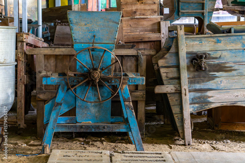 Old blue agricultural threshing machine with hand wheel among wooden crates in a rustic farm setting.