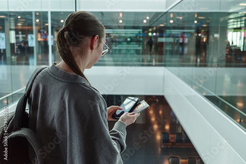 Young woman with a backpack holding a passport and smartphone at a contemporary airport. Concept of international travel, identity verification and mobility.