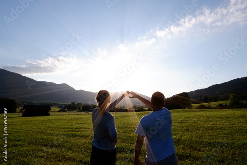 Paar formt Herz mit Händen vor Sonnenuntergang in grüner Berglandschaft