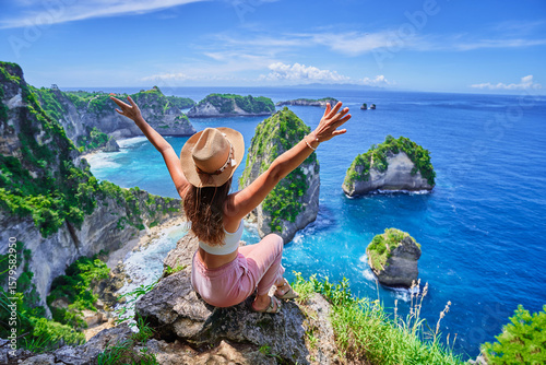 Woman traveler with open arms sitting on rock cliff and enjoying of scenic landscape of Diamond beach during vacation to Nusa Penida Island in Indonesia, travel to touristic beautiful destinations
