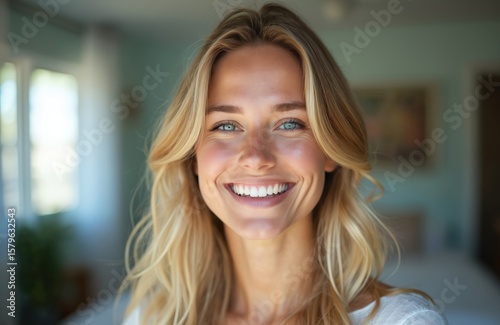 Cheerfull blonde woman smiling brightly with turquoise eyes in cozy interior room wearing white top. Long blonde hair frames face, showing natural beauty, confidence. Portrait captures joy, positive
