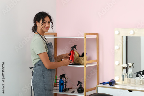 Slika na platnu Beautiful young happy African-American hairdresser taking spray from shelving un