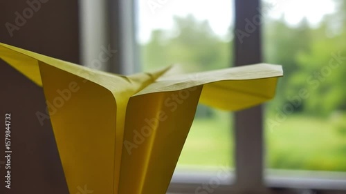 Close up shot of a yellow paper airplane with a window and greenery in the background indoors