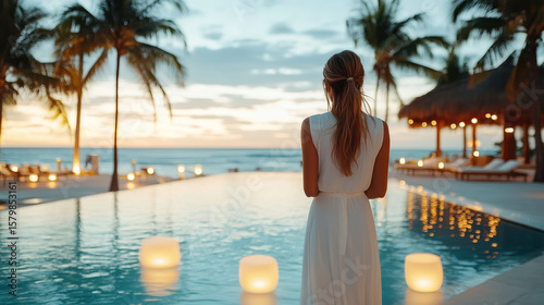 Elegant woman white dress standing by resort pool sunset, surrounded by palm trees and lights