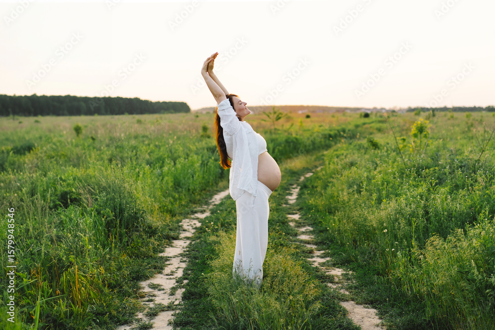 Fototapeta premium A pregnant woman stands in a green field. She gazes lovingly at her baby bump as the sun sets behind her, creating a serene atmosphere.
