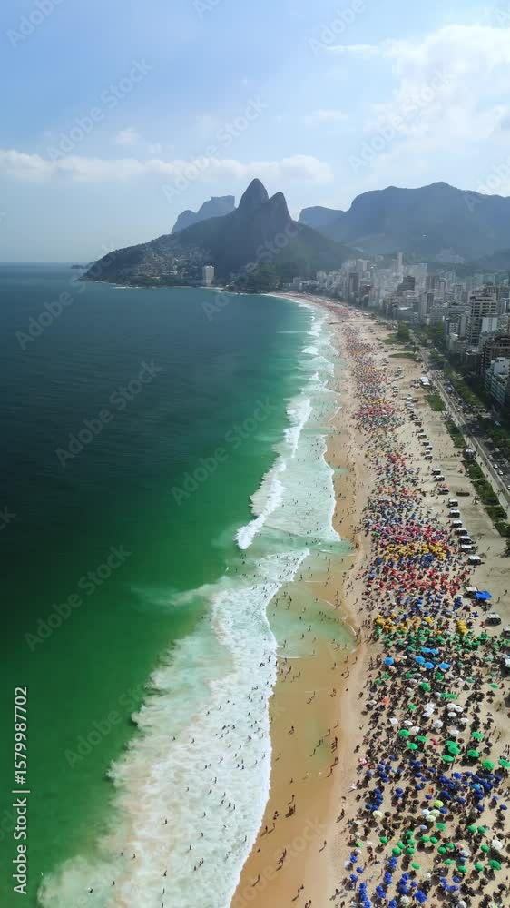 Vertical drone flyover shows Ipanema’s umbrella-packed beach, emerald surf, Dois Irmãos